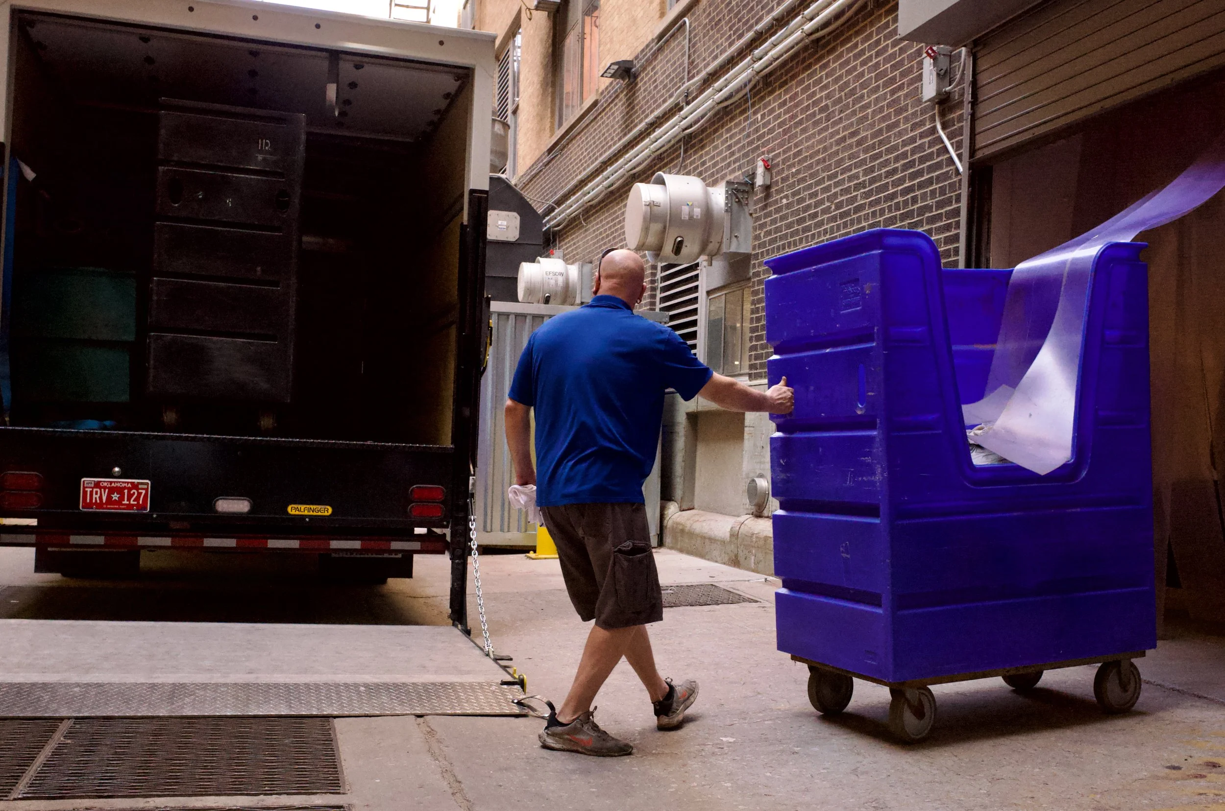 A man in a blue shirt and shorts pulling a blue recycling cart in an alleyway near a truck.