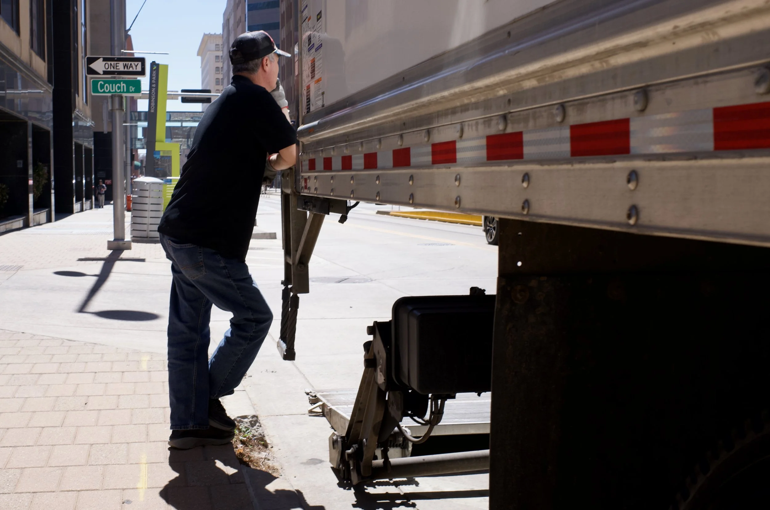 A man in a black t-shirt, jeans, and a cap is guiding or inspecting the side of a large truck that is parked on a city sidewalk. Streets signs visible include Couch Drive and Robinson Plaza, indicating an urban environment.