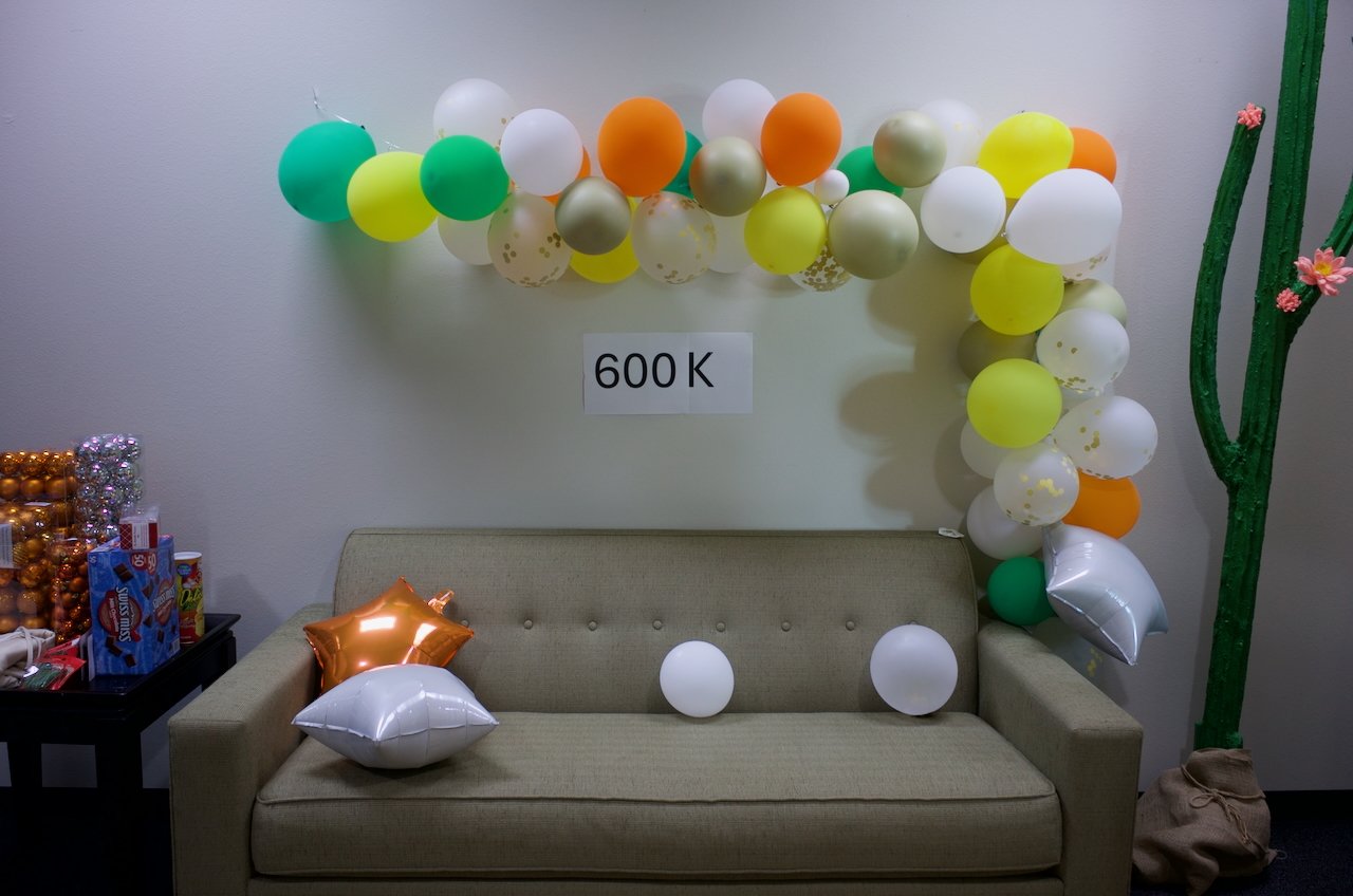 Decorated room with a beige sofa, a balloon arch on the wall including green, yellow, white, orange, and gold balloons, and a sign that reads '600 K'. There are star-shaped balloons on the sofa, and on the left, there is a table with party supplies a