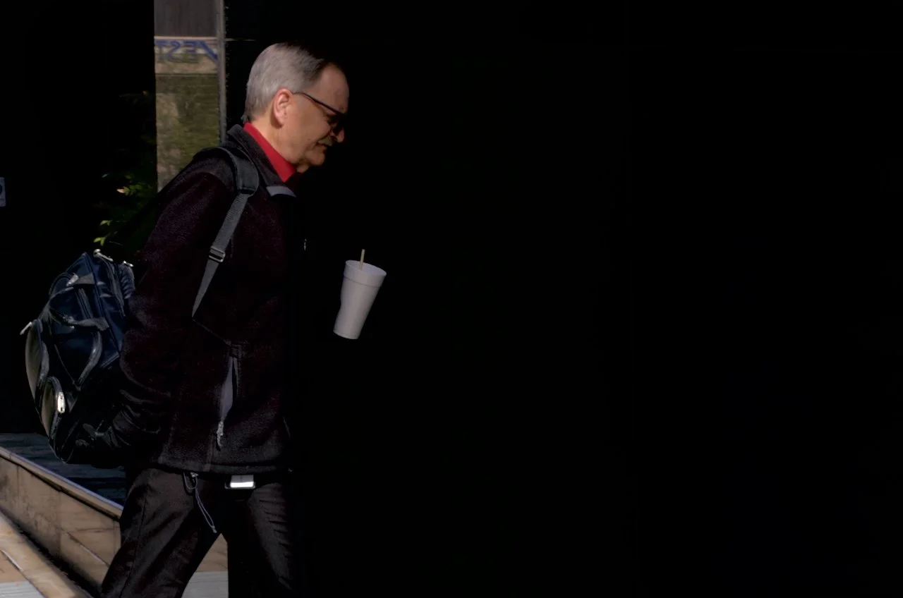 A man with gray hair wearing glasses, a black jacket, red shirt, holding a white styrofoam cup with a straw, and carrying a backpack, walking outdoors at night.