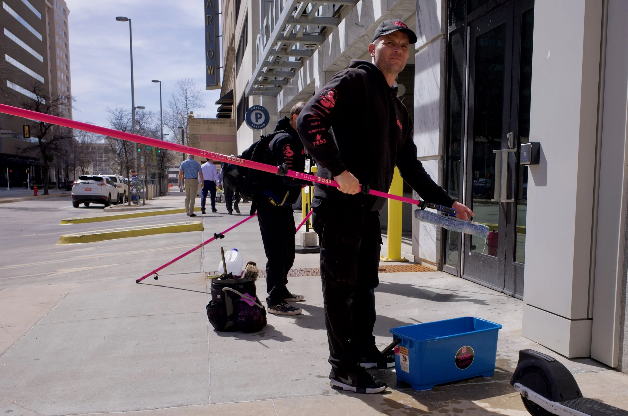 A man cleaning sidewalk at a storefront entrance with pink broom, surrounded by cleaning supplies, while others in black attire work behind him. Urban street scene with buildings, parked cars, and pedestrians.
