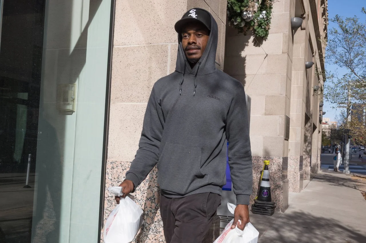 A man in a gray hoodie and black cap walking on a city sidewalk, holding plastic bags in both hands. There are trees, buildings, and a crosswalk in the background.