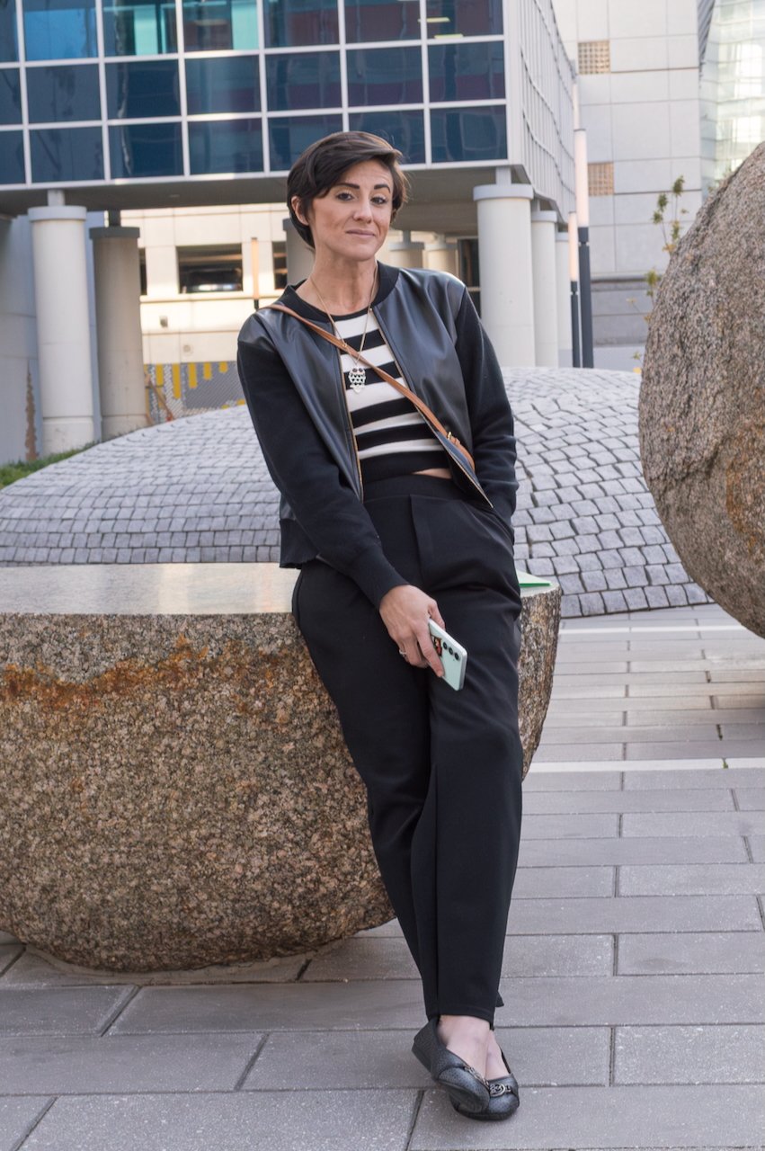 A woman with short brown hair leaning against a rock sculpture in an urban outdoor setting, holding a smartphone, wearing a black leather jacket over a black and white striped crop top, black pants, and black flats.