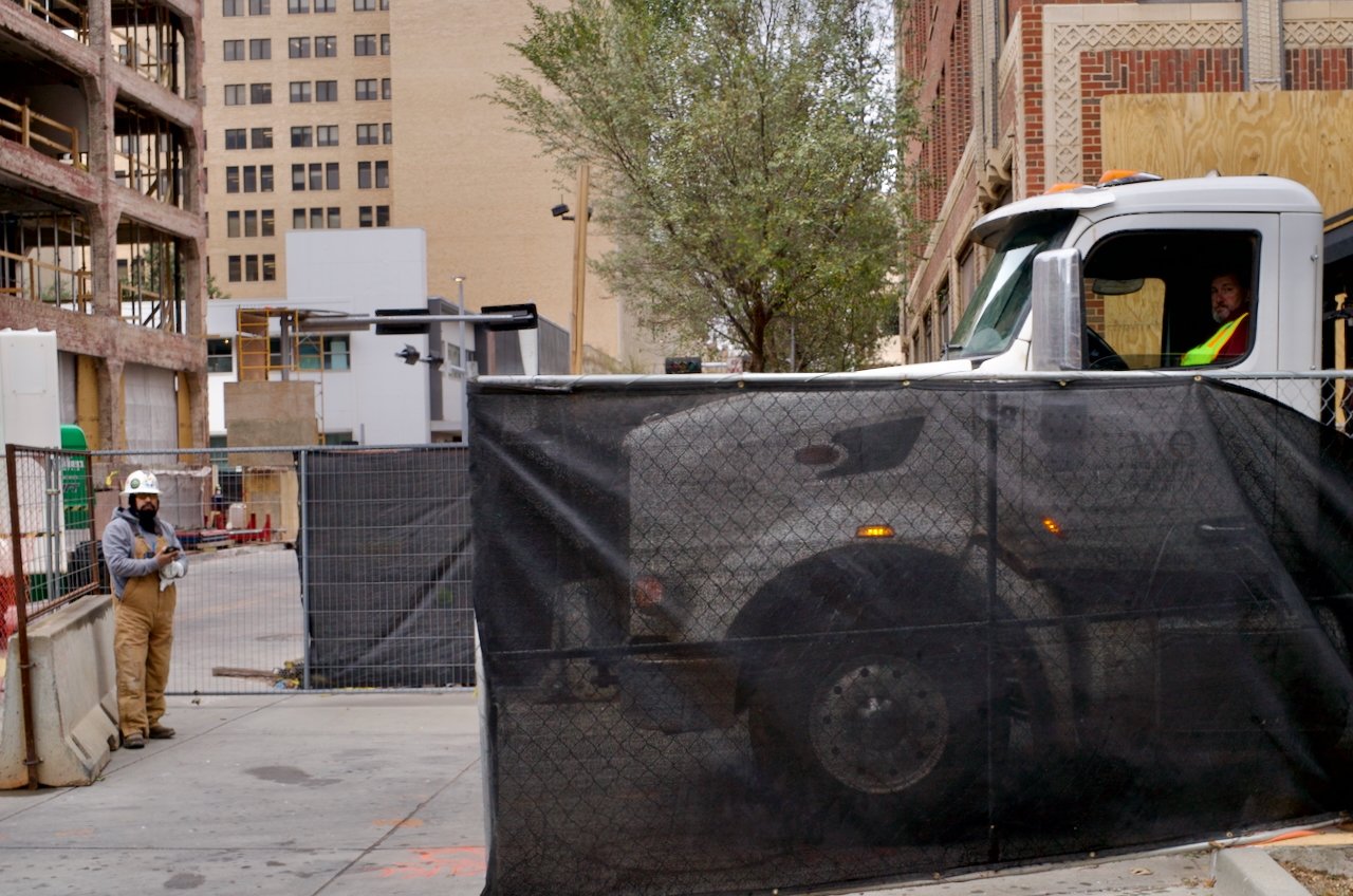Construction site with workers and building materials, one person in a hard hat and safety gear standing near a black fence, and a white truck parked close to a building under construction with a person visible in the driver's seat.