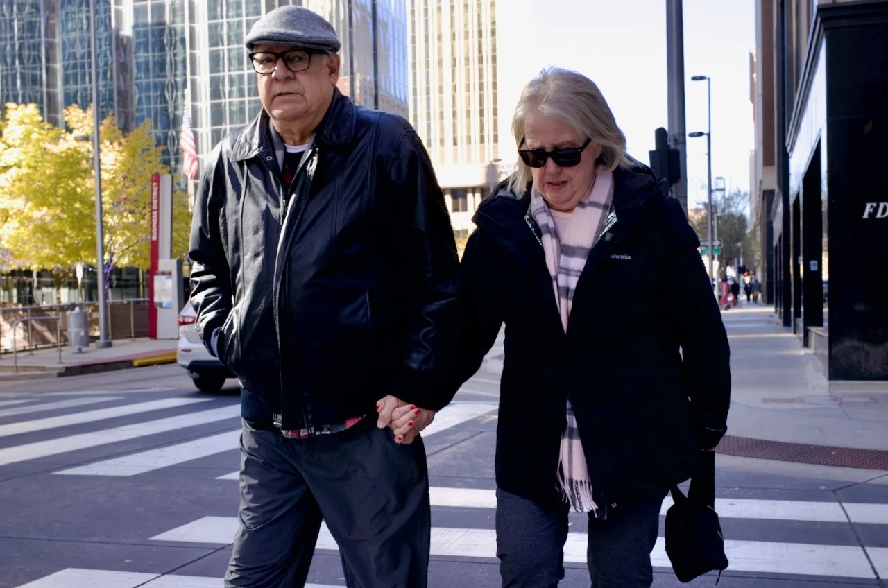 A mature man and woman walking hand in hand across a city crosswalk on a sunny day, wearing jackets and sunglasses.