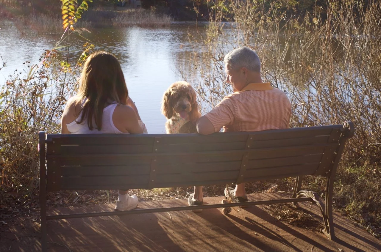 A girl, an older man, and a dog sitting on a bench by a pond, enjoying nature during autumn.