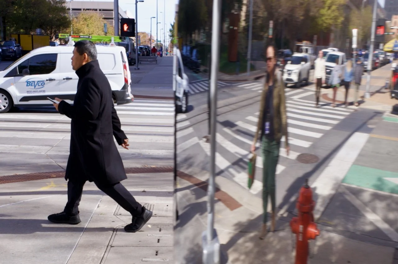 Two pedestrians walking on downtown OKC sidewalks, one man in a black coat looking at his phone crossing the street, and a woman carrying a bag walking behind him. Cars parked along the street and traffic signals visible.