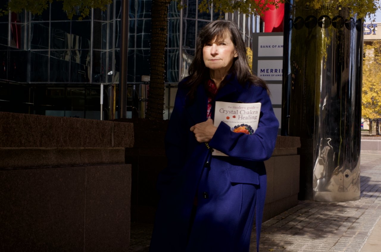 A woman with long dark hair stands outdoors holding a book titled "The Modern Guide to Crystal Chakra Healing." She is wearing a navy blue coat and has a serious expression, with trees and modern buildings in the background.