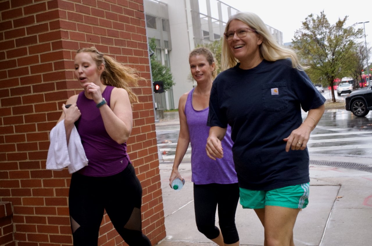 Three women running outdoors on a city sidewalk during a rainy day, all dressed in athletic clothing.