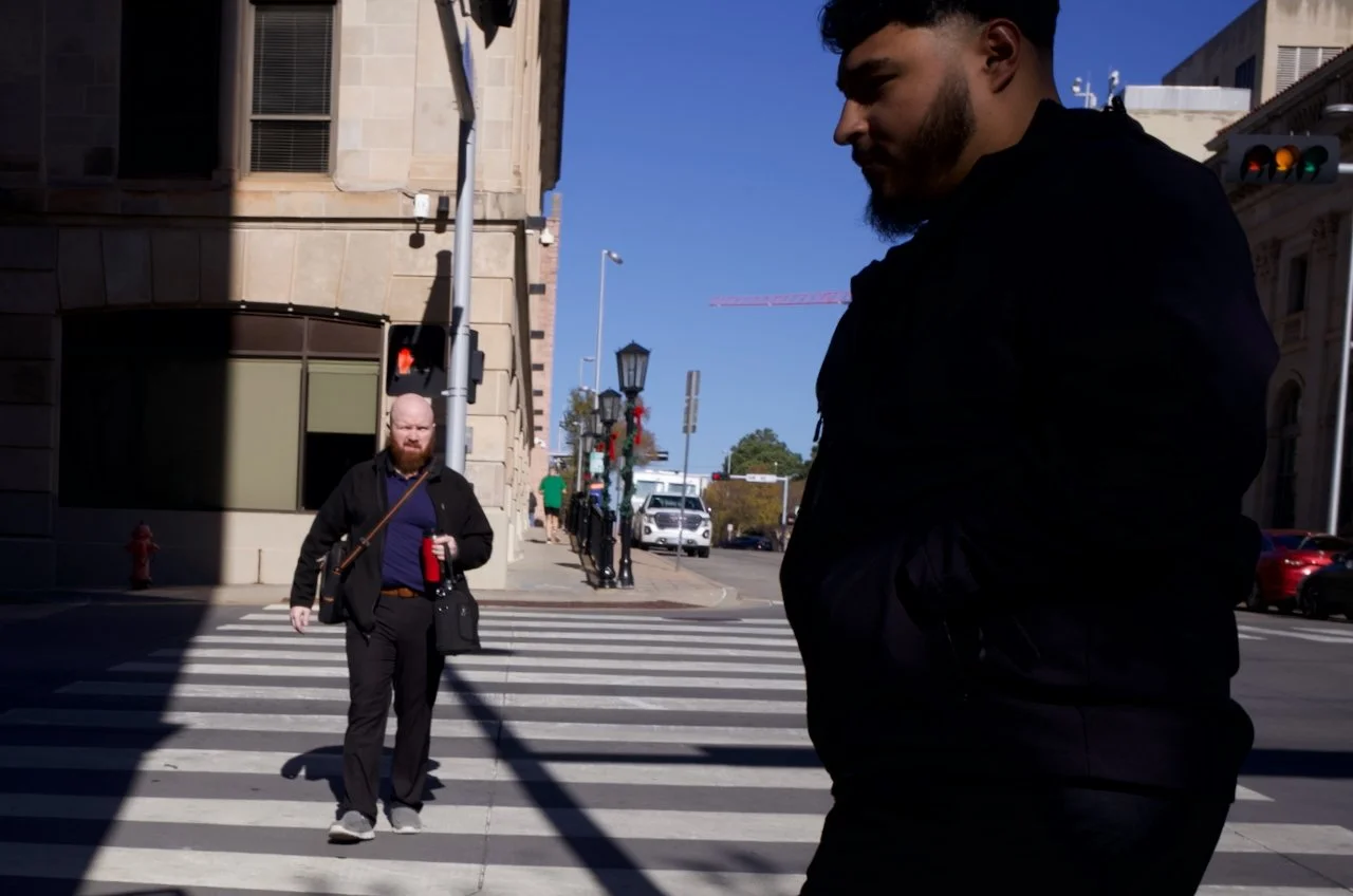 Two men crossing a city street at a crosswalk with traffic lights, one man is on the right side of the image, and the other is in the background holding a drink, with a building and cars in the background.