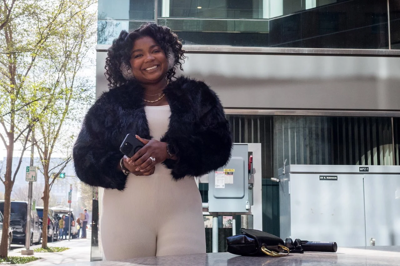 Smiling woman with curly hair wearing a black fur coat and white outfit, holding a phone, standing outside with trees and buildings in the background.