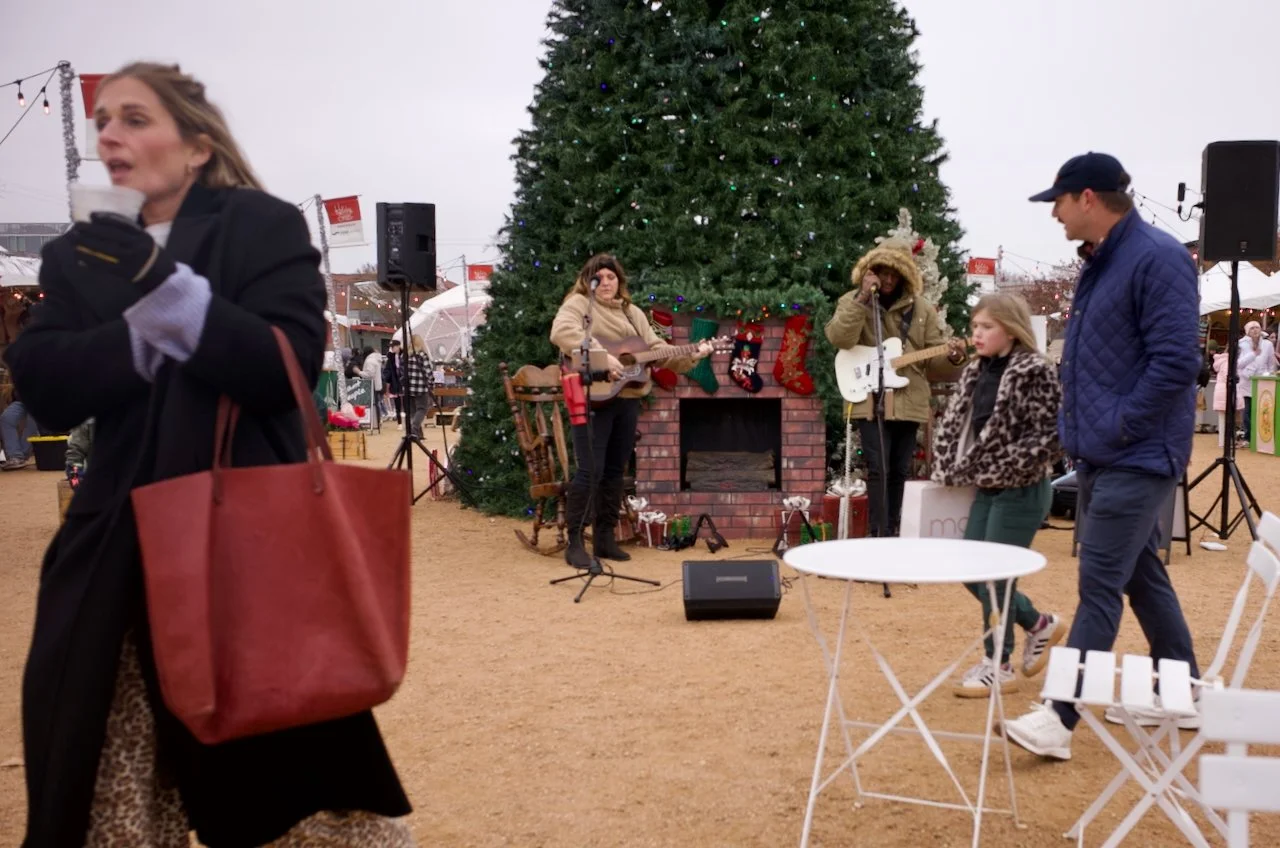 People gathered around a large decorated Christmas tree at an outdoor festival, with musicians performing near the tree, and a woman in the foreground holding a red tote bag and a beverage.