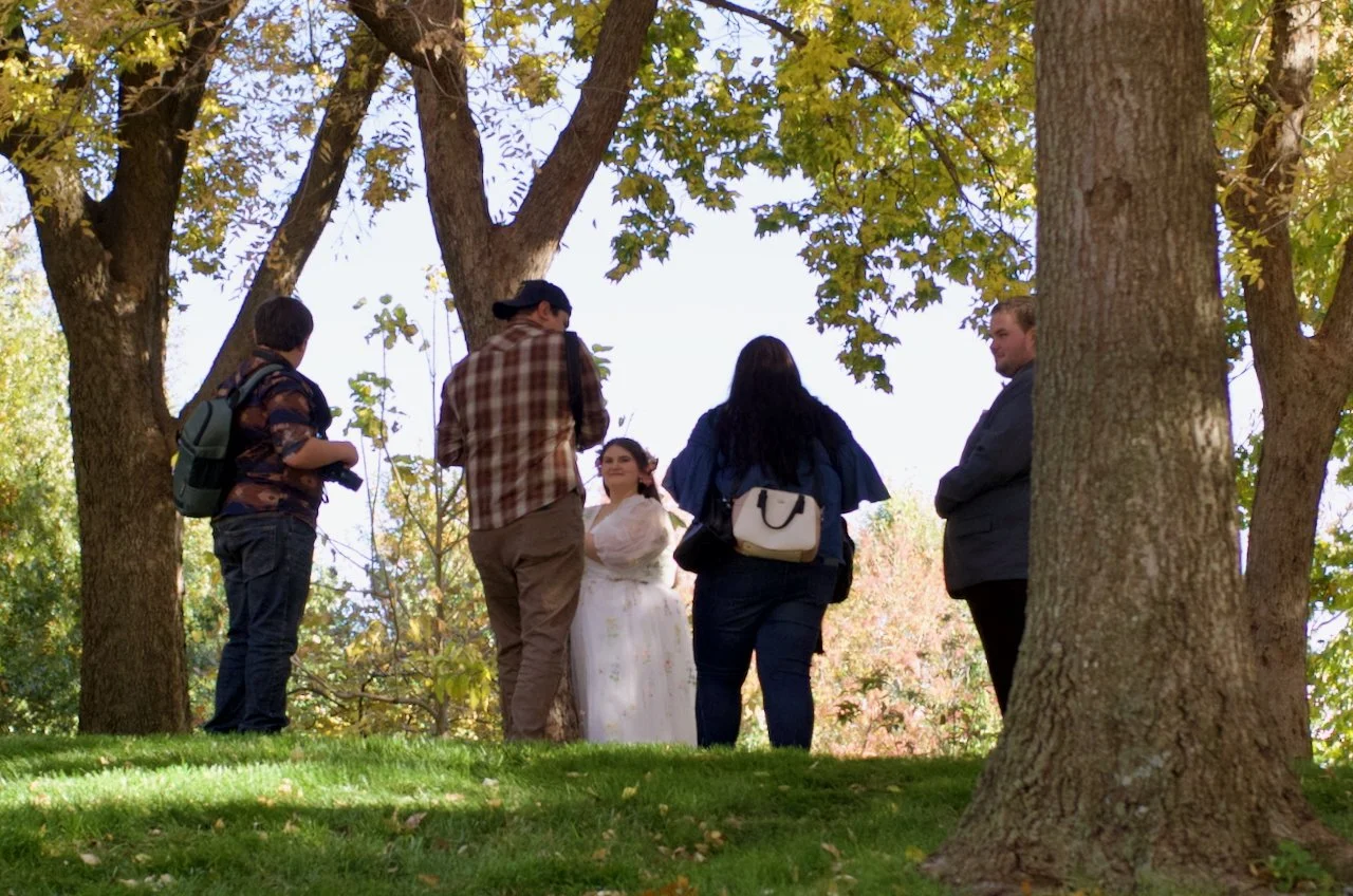 People gathered outdoors under trees, with one woman in a white dress, possibly for a photoshoot or event.