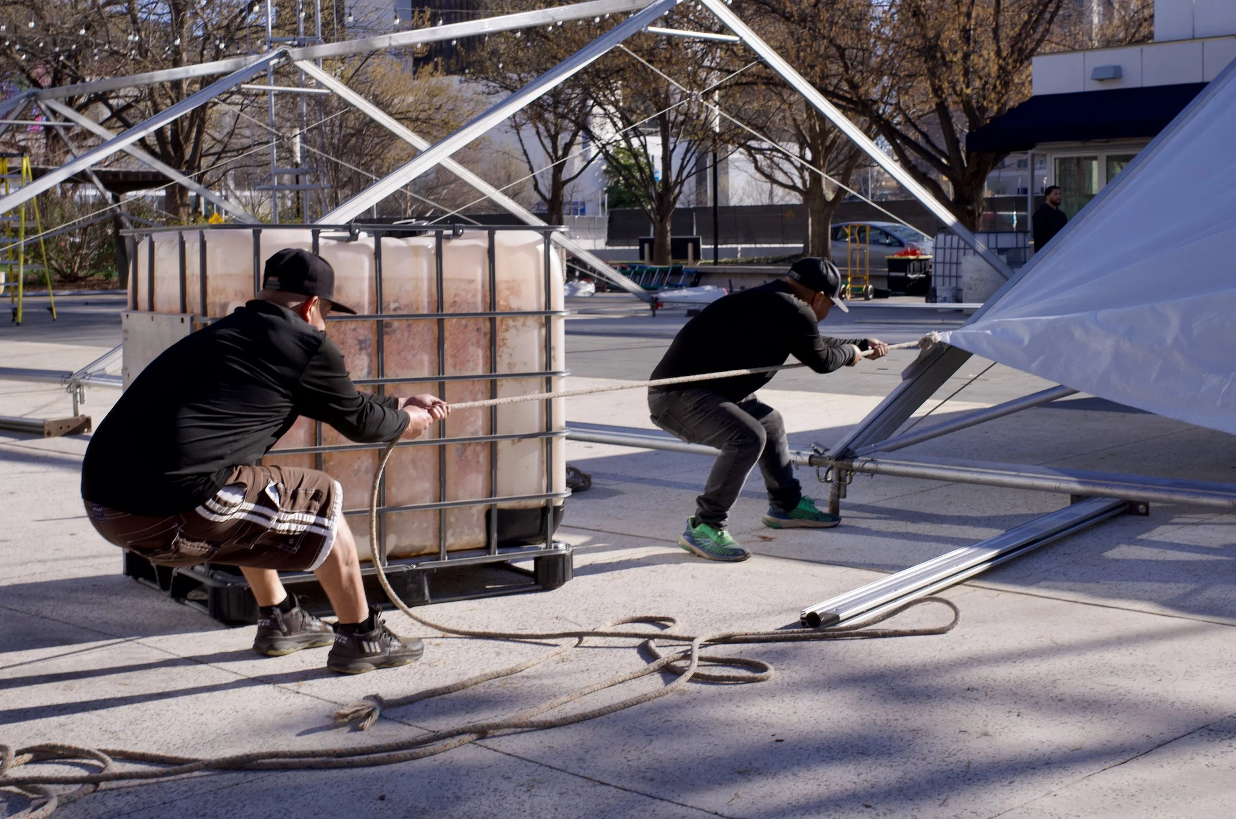 Two men are pulling down a large metal frame structure while working on assembling or disassembling it outside.