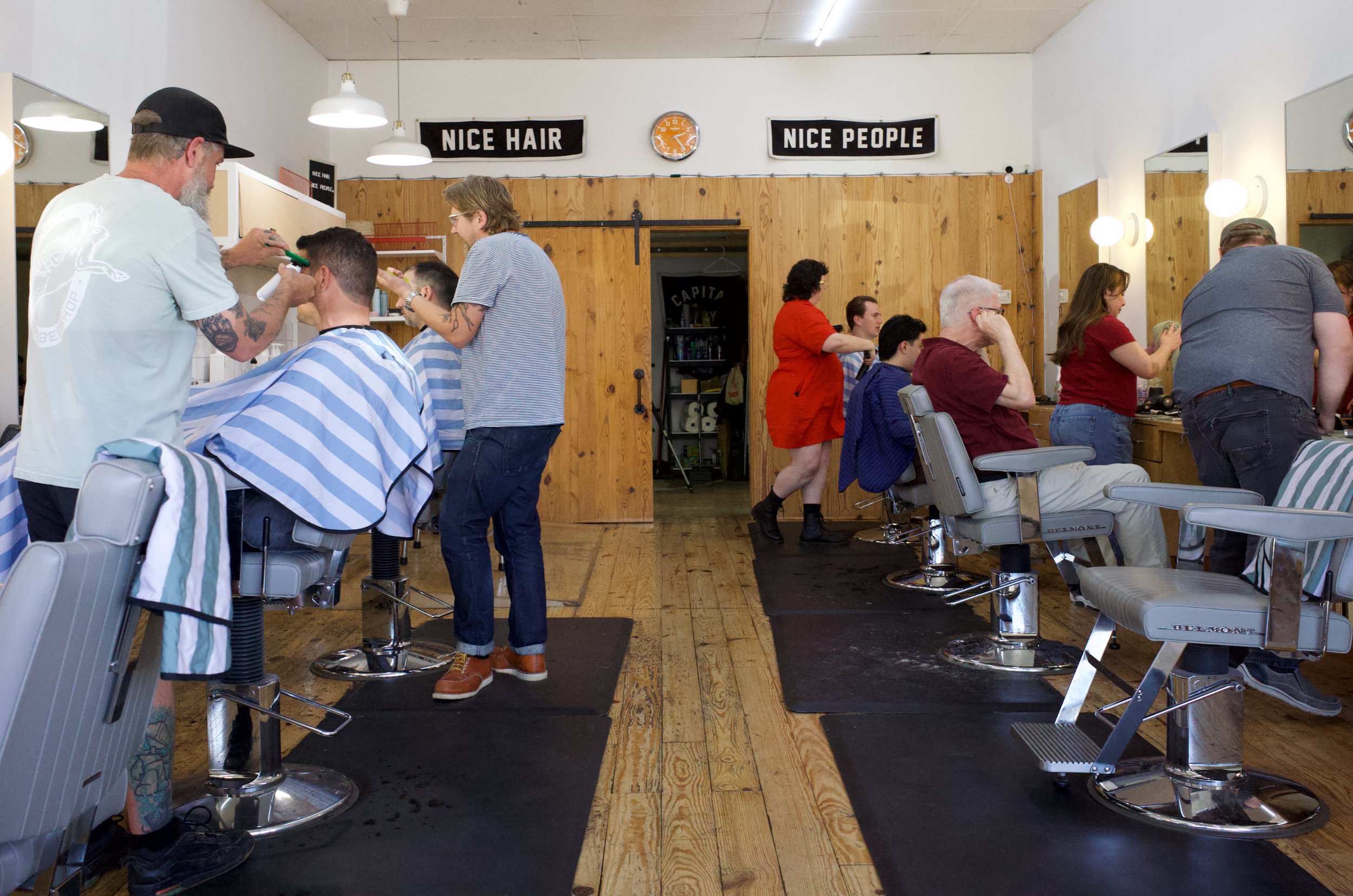 Inside a barbershop with customers getting haircuts and stylists working. Signage reads 'NICE HAIR' and 'NICE PEOPLE'.