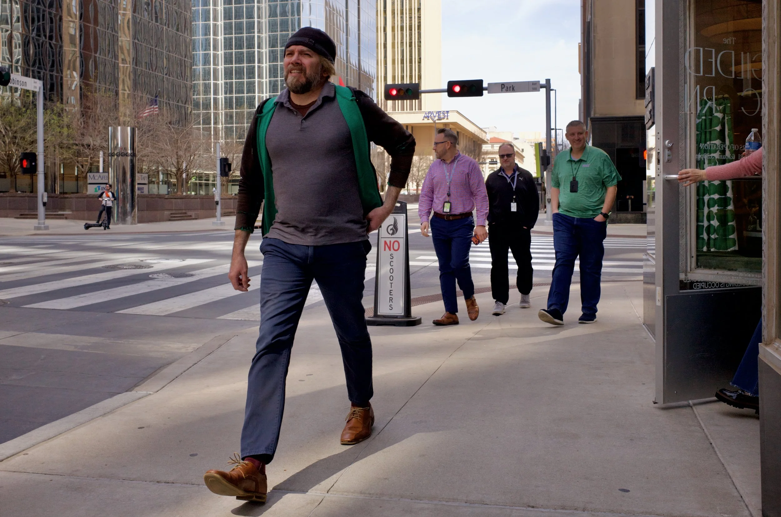 A man with a beard, wearing a dark beanie, gray shirt, and brown shoes, walking on a city sidewalk with three other men in the background. They are near a crosswalk at an urban intersection, with tall buildings and traffic lights visible.