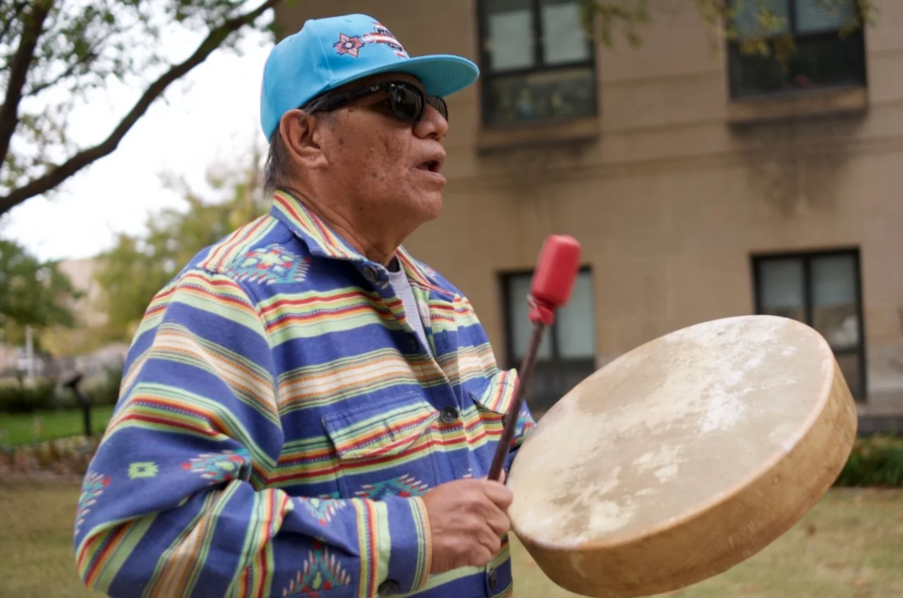 A man wearing a colorful striped jacket, a blue cap, and sunglasses is playing a large musical drum outdoors.