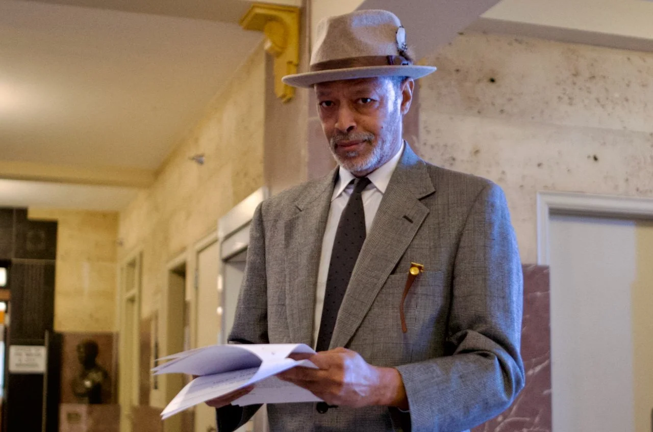 OKC's Michael Washington wearing a gray plaid suit, white shirt, black tie, and beige fedora hat with a ribbon, holding documents in Oklahoma City City Hall.