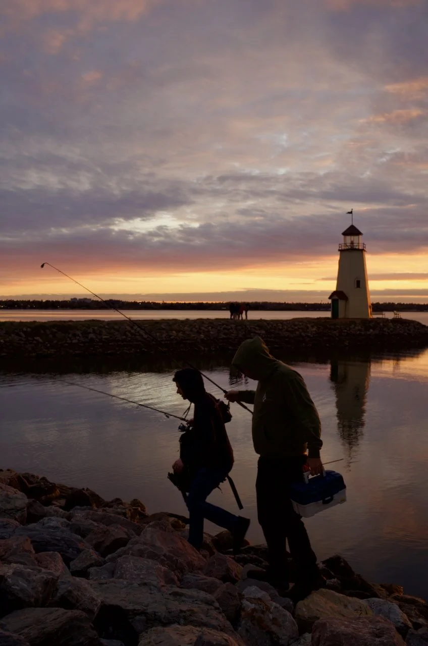 Two people are walking along a rocky shoreline at sunset, carrying fishing rods. A lighthouse stands in the distance near a body of water, with a few horses visible further away. The sky is partly cloudy with vibrant sunset colors.