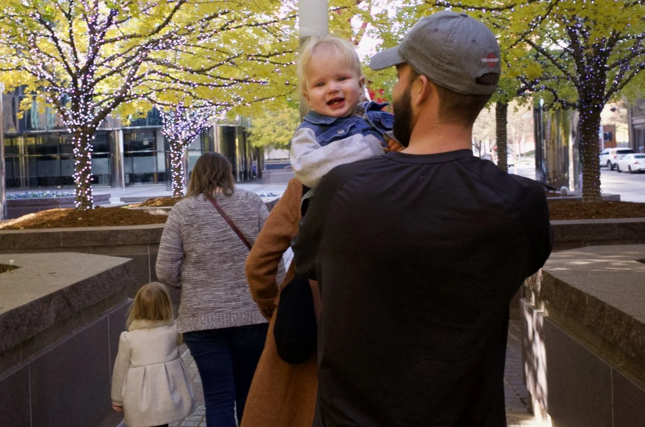 A man holding a happy young boy outside near trees decorated with string lights, with other pedestrians walking in the background.