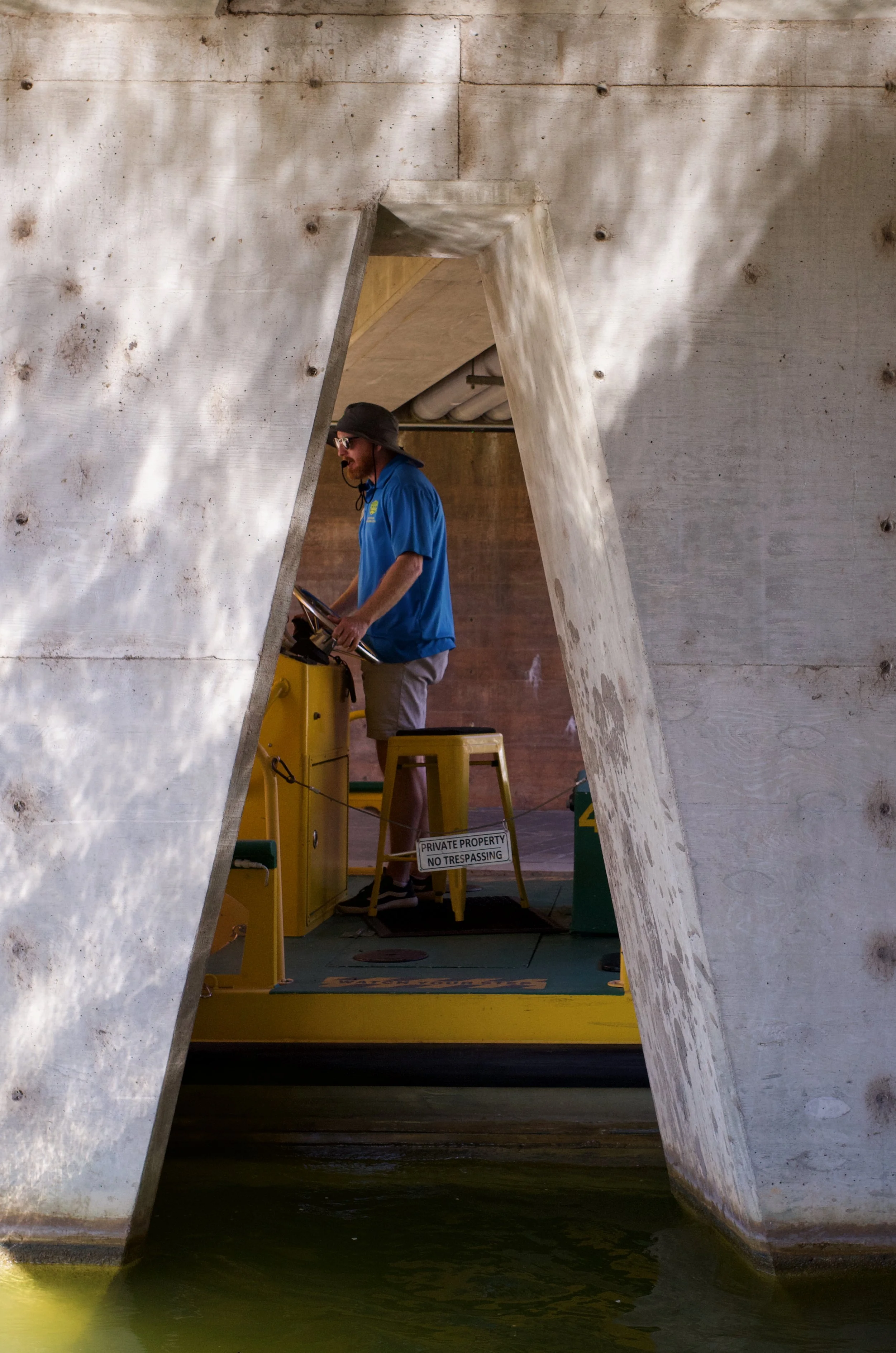 A man with sunglasses and a hat using a control panel in a structure with concrete walls, seen through a triangular opening, next to water.