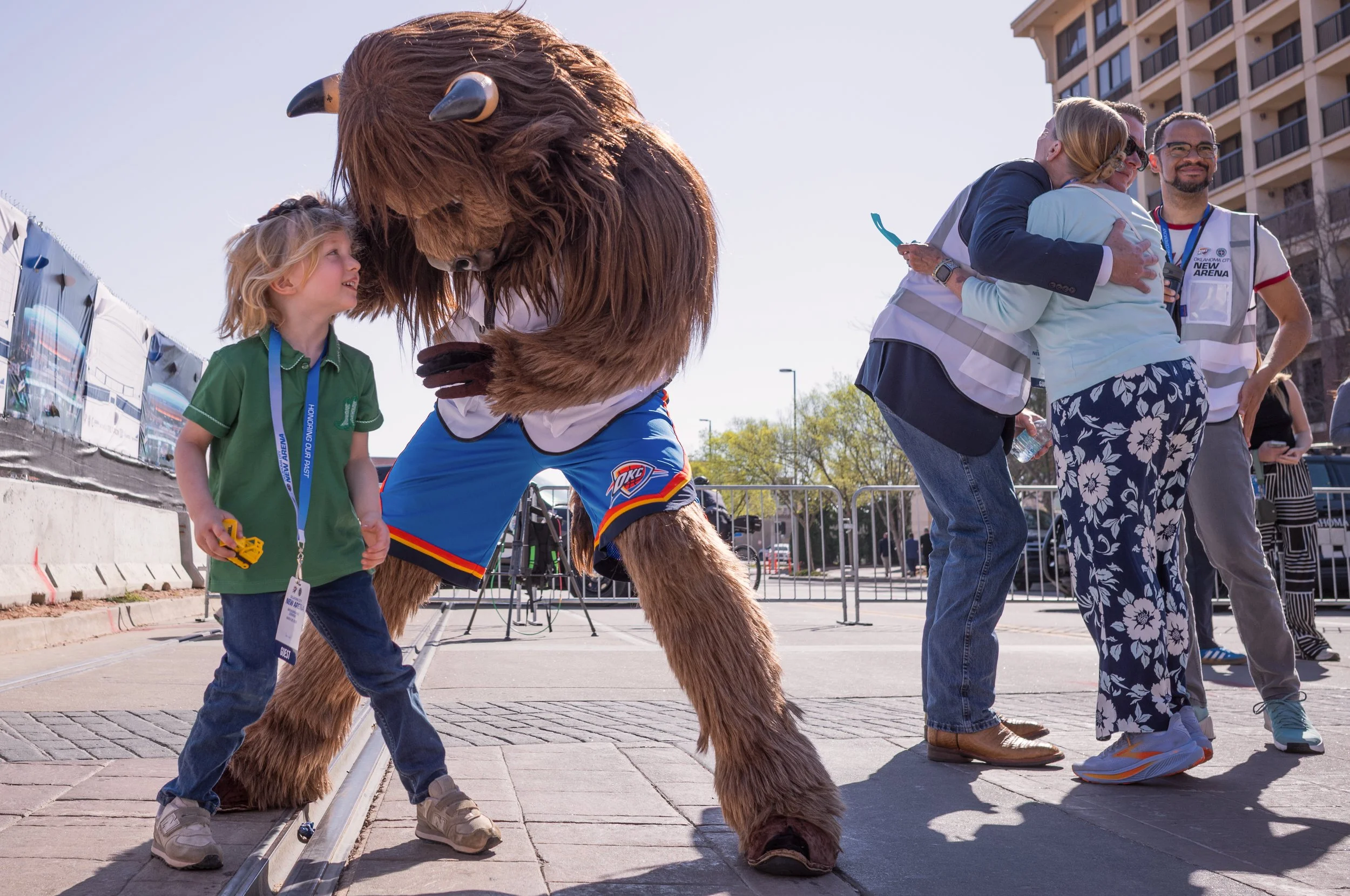 A young child and a person in a mascot costume of a bear character with basketball shorts are interacting outdoors, with onlookers in the background.