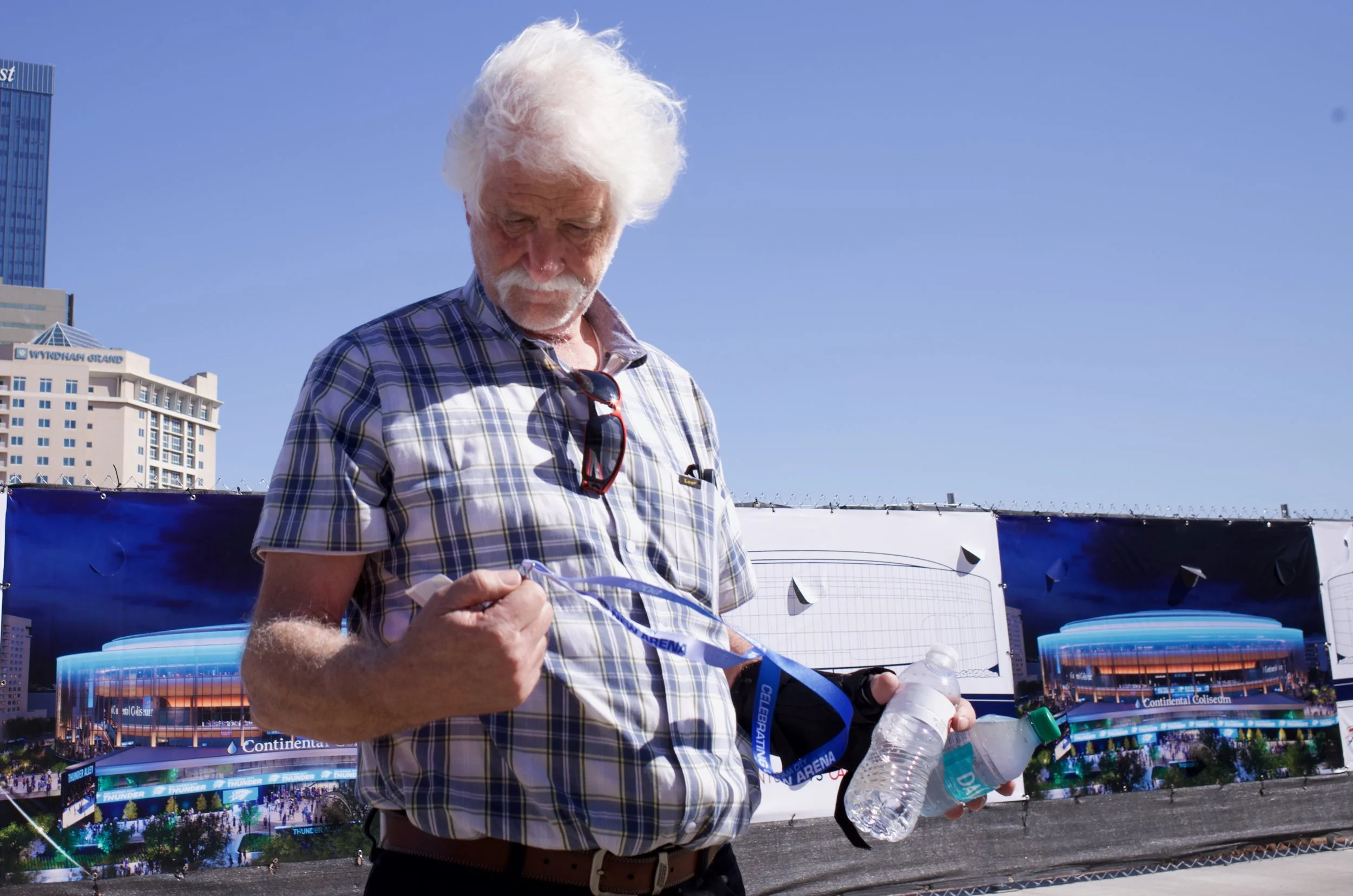 Older man with white hair and beard wearing a plaid shirt, holding bottled water, standing outdoors near a banner with an architectural rendering of a stadium, under a clear blue sky.