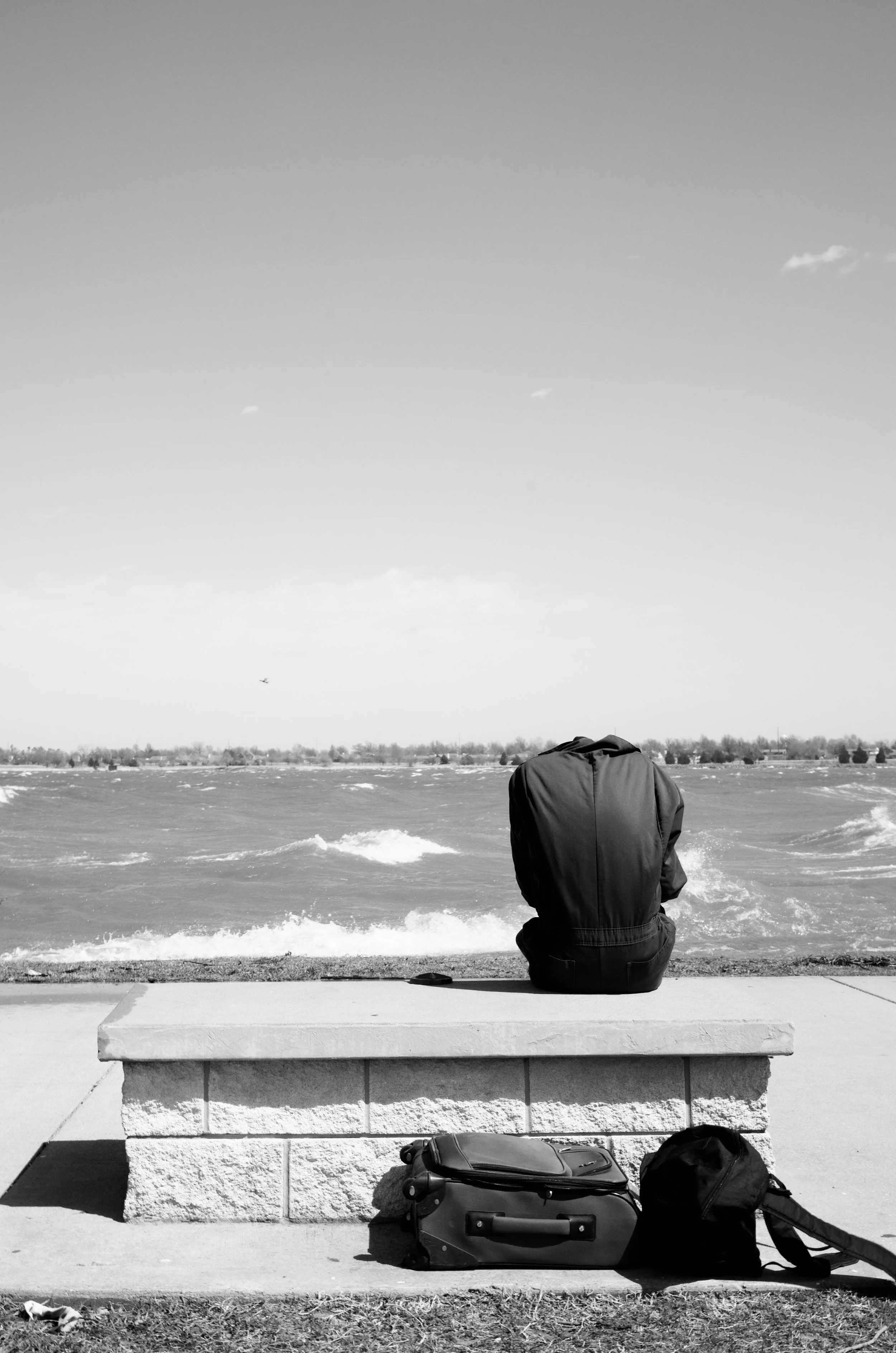 A person sitting on a stone bench by the water, with luggage and a backpack on the ground in front of them, gazing at the river on a clear day. This photo, Rough Waters, was selected as a Leica Master Shot by LFI – Leica Fotografie International.