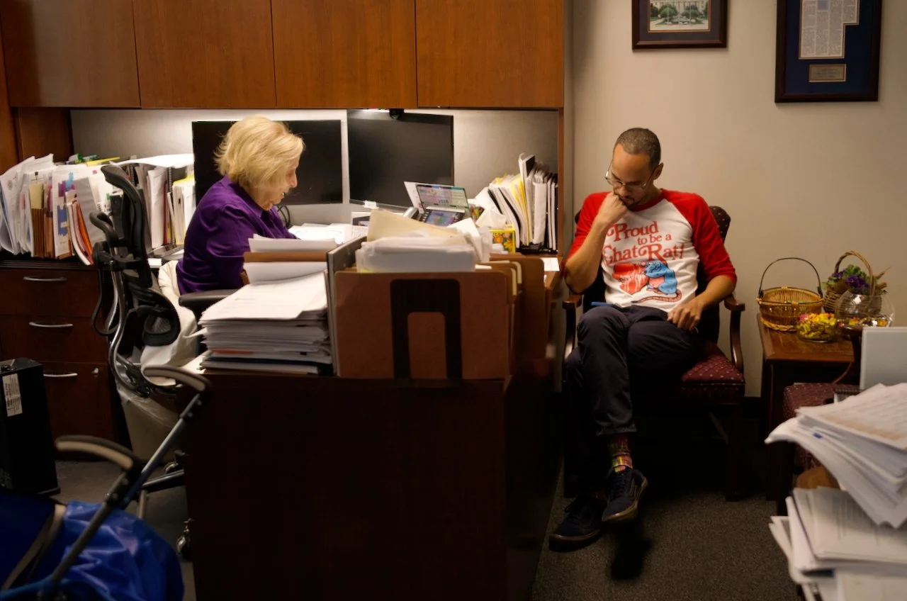 An office with a woman working at her desk and a man sitting on a chair. The woman has blond hair and is wearing a purple shirt. The man has short hair, glasses, a red and white shirt, and colorful socks. There are multiple computer monitors, paperwo