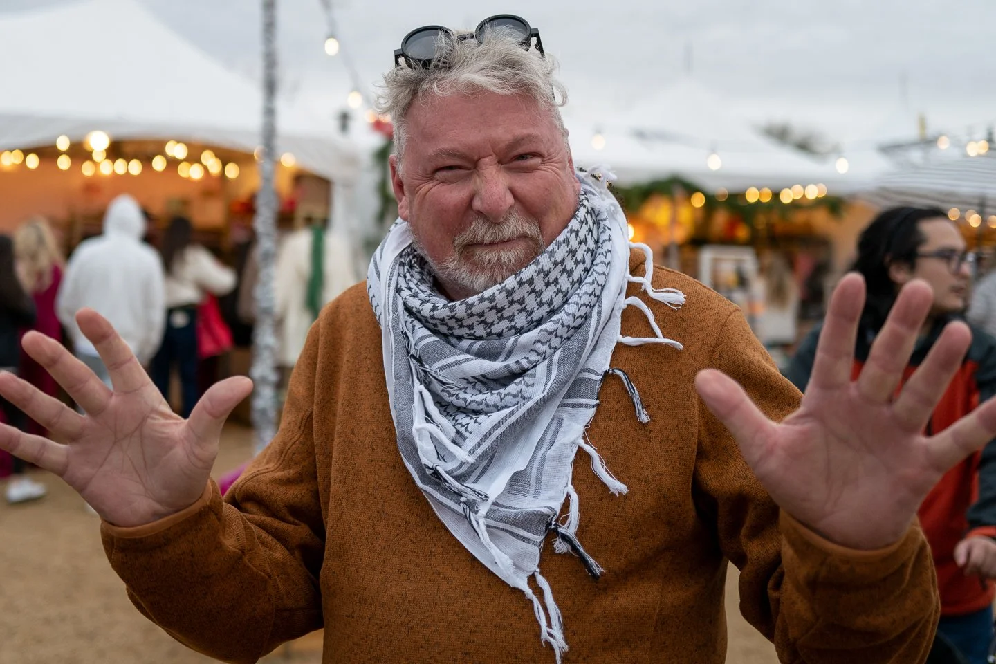 A smiling man with gray hair and beard, wearing sunglasses on his head, a white and black patterned scarf, and a brown jacket, at an outdoor event with tents and lights, spreading his hands toward the camera.