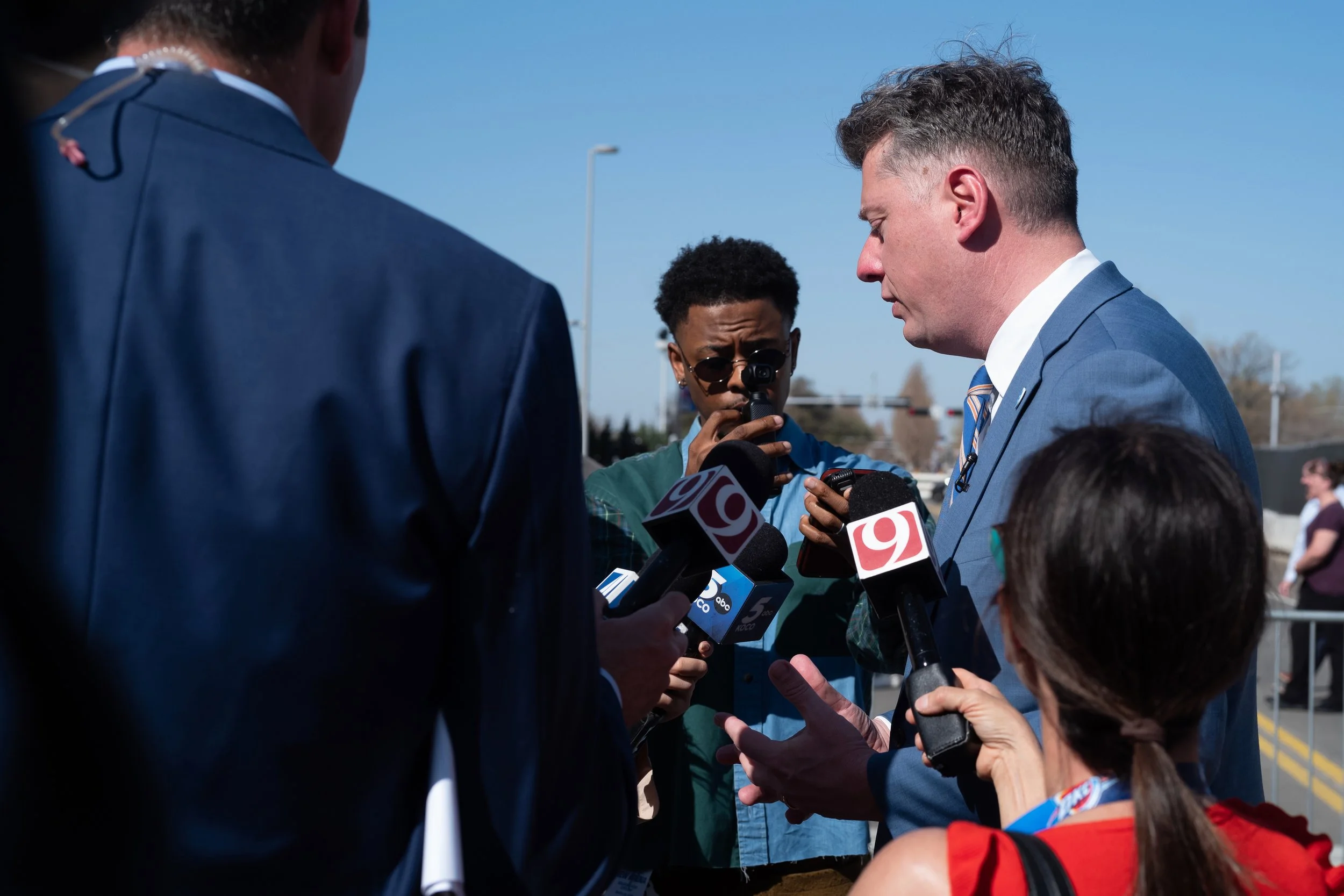 A man in a blue suit giving a statement to reporters holding microphones with news station logos, while a woman with sunglasses and a man with glasses and a camera observe.
