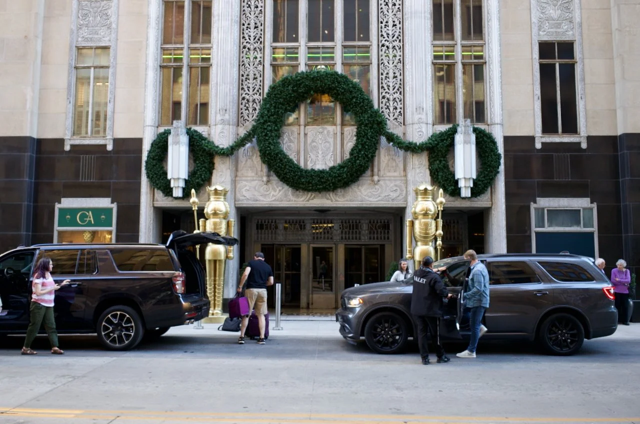 People standing near two black SUVs outside a building decorated with a large green wreath and gold nutcracker decorations for Christmas.
