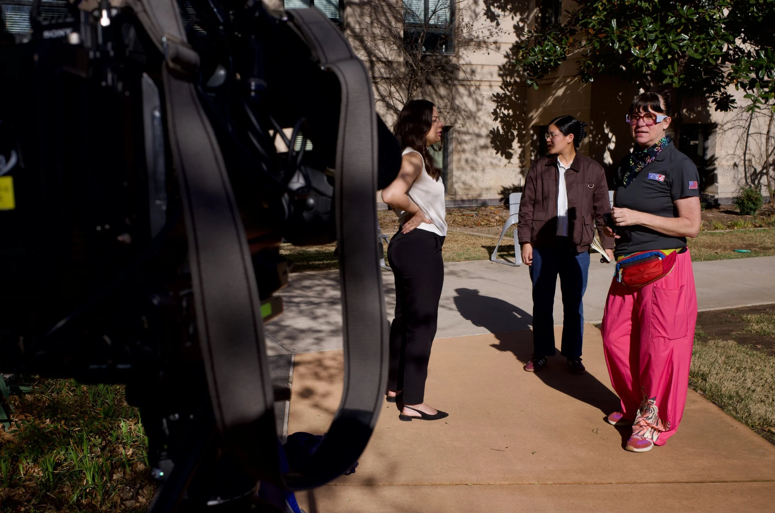 Three women engaged in conversation outdoors, with a camera recording the scene. One woman is wearing a white top and black pants, another is wearing glasses and a brown jacket, and the third woman has glasses, a black polo shirt, and pink pants.
