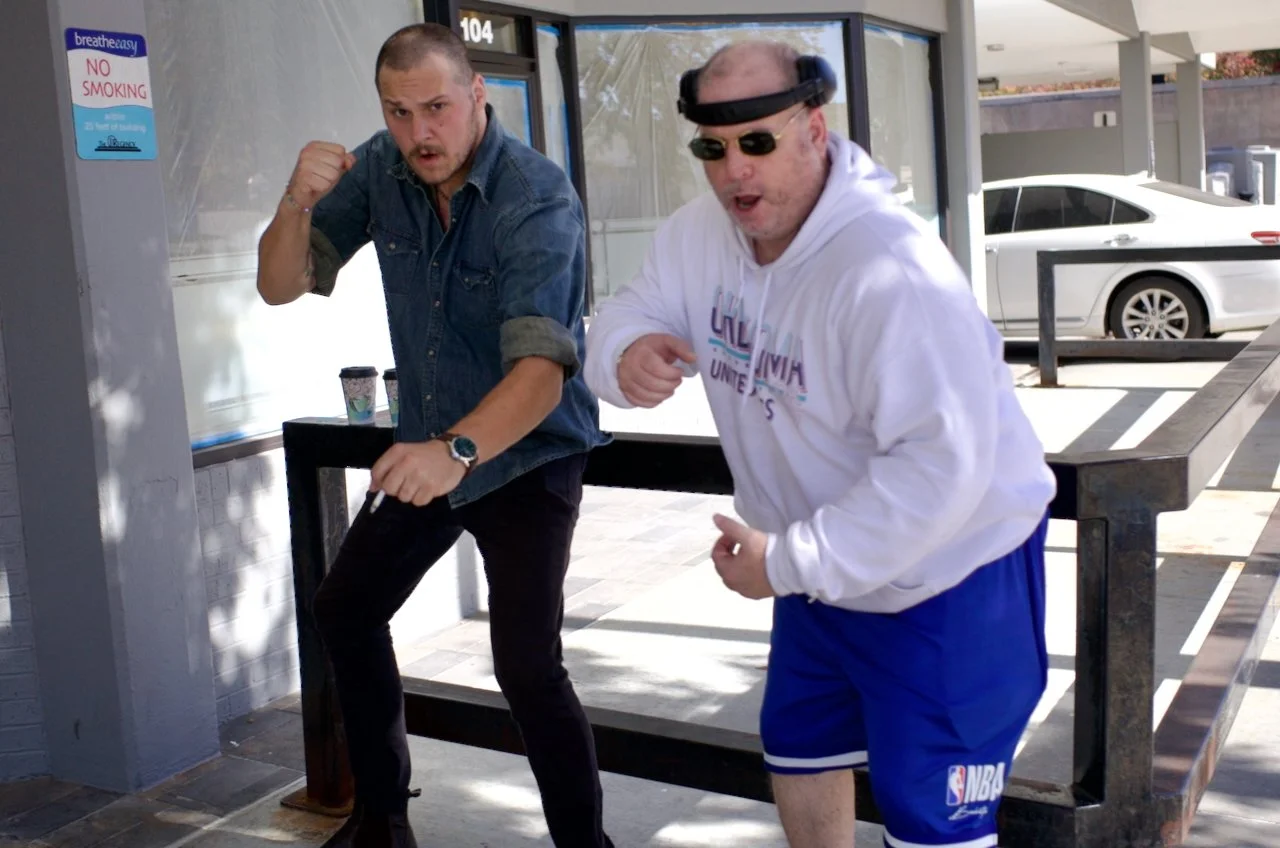 Two men standing outside a building, one in a denim shirt and the other in a white hoodie and blue shorts, both making fighting poses. A parked car and a 'No Smoking' sign are visible in the background.