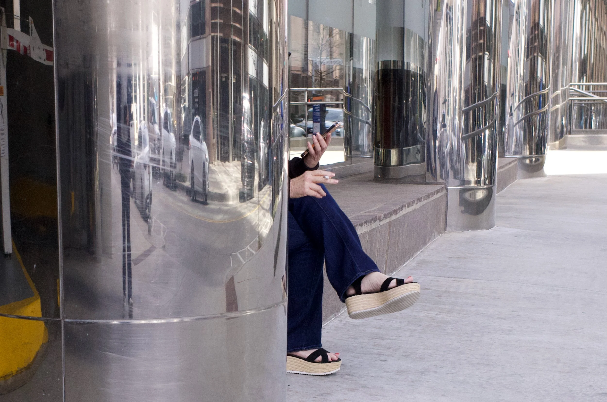 A person sitting on a brick ledge outside a building, using a cellphone, with their face obscured by a reflective metal pillar.