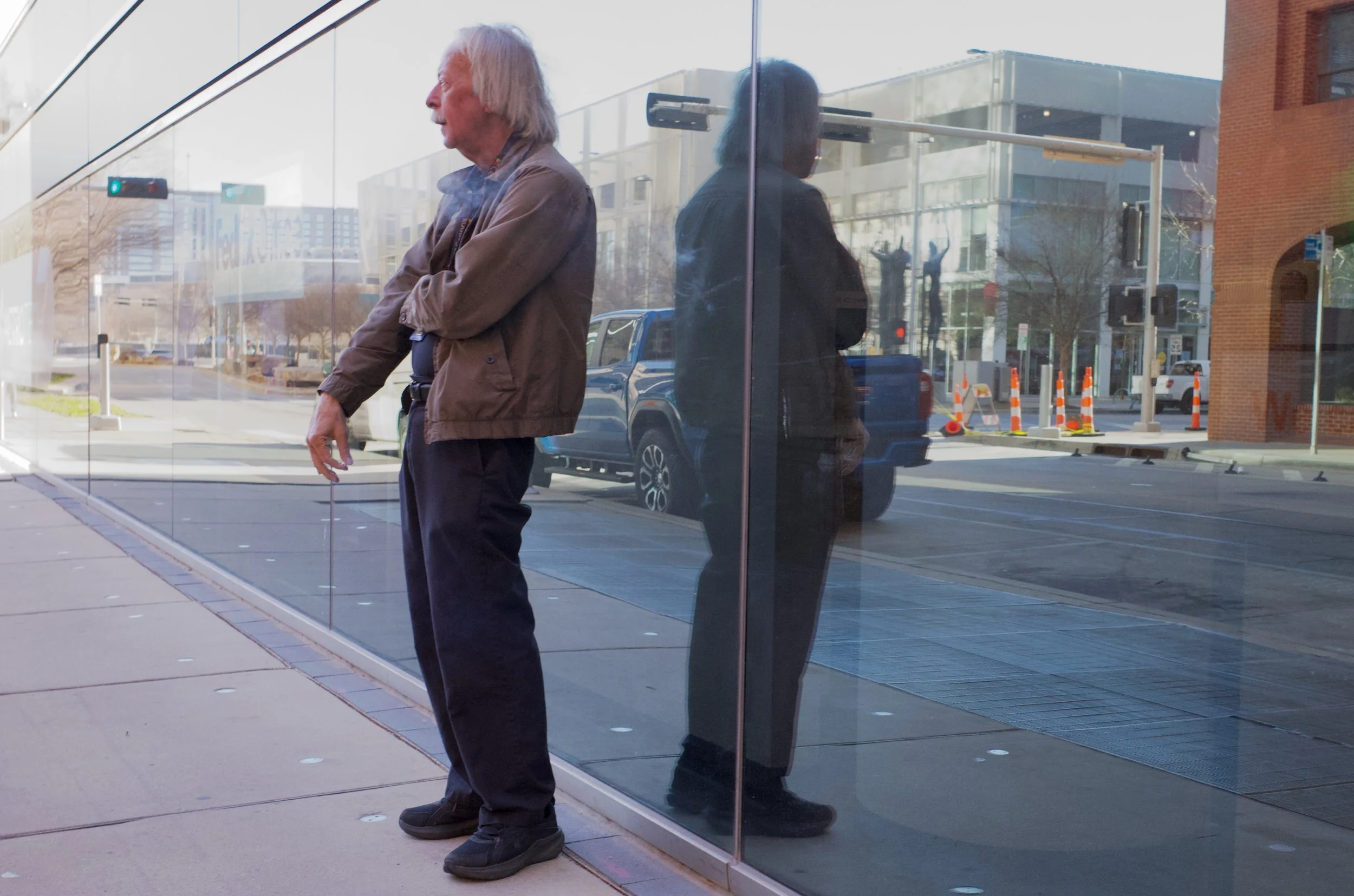 An older man with white hair, wearing a brown jacket and black pants, leaning against a glass wall of a building. His reflection is visible in the glass, and he stands outside on the sidewalk.