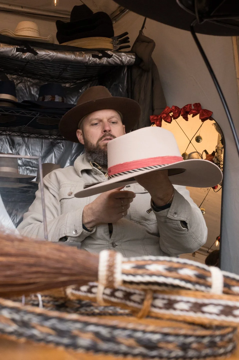 Man wearing a cowboy hat and beige shirt holding a white hat with red band indoors.