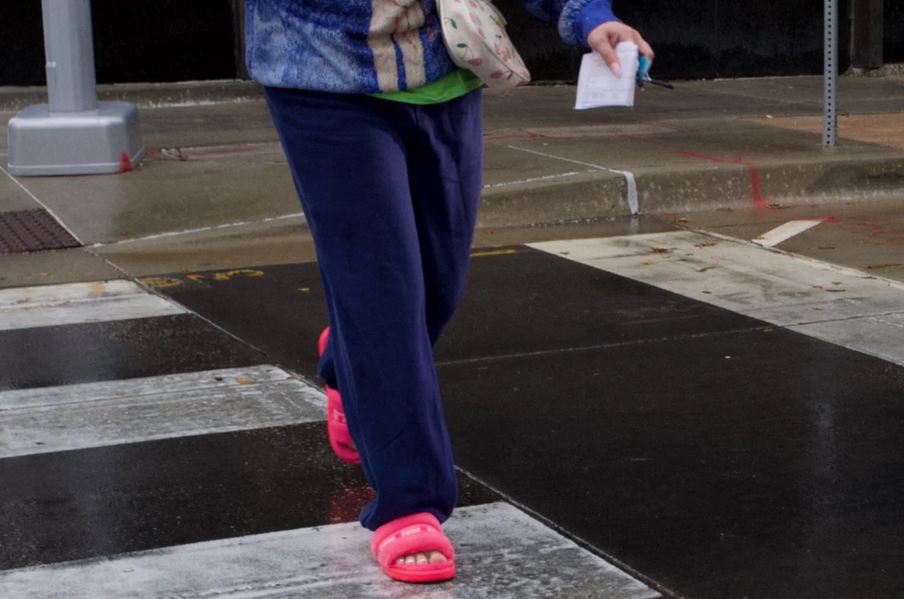 Person crossing a pedestrian crosswalk on a street, wearing blue pants and bright pink slides, holding a piece of paper and other items in their hand.