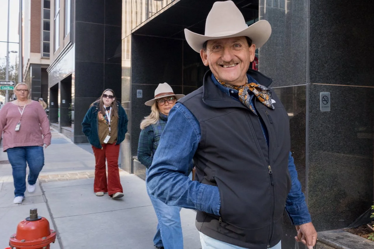 A smiling man wearing a cowboy hat, denim shirt, vest, and scarf standing on a city sidewalk with three women in casual clothing walking behind him.