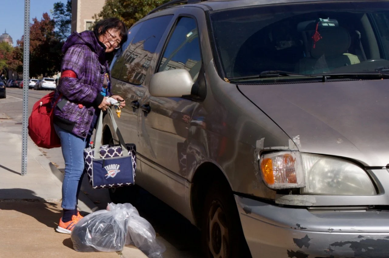 Woman with glasses, purple plaid jacket, and red backpack on sidewalk, holding a bag of trash, next to a damaged silver minivan with a missing headlight, on a city street with trees and buildings in the background.