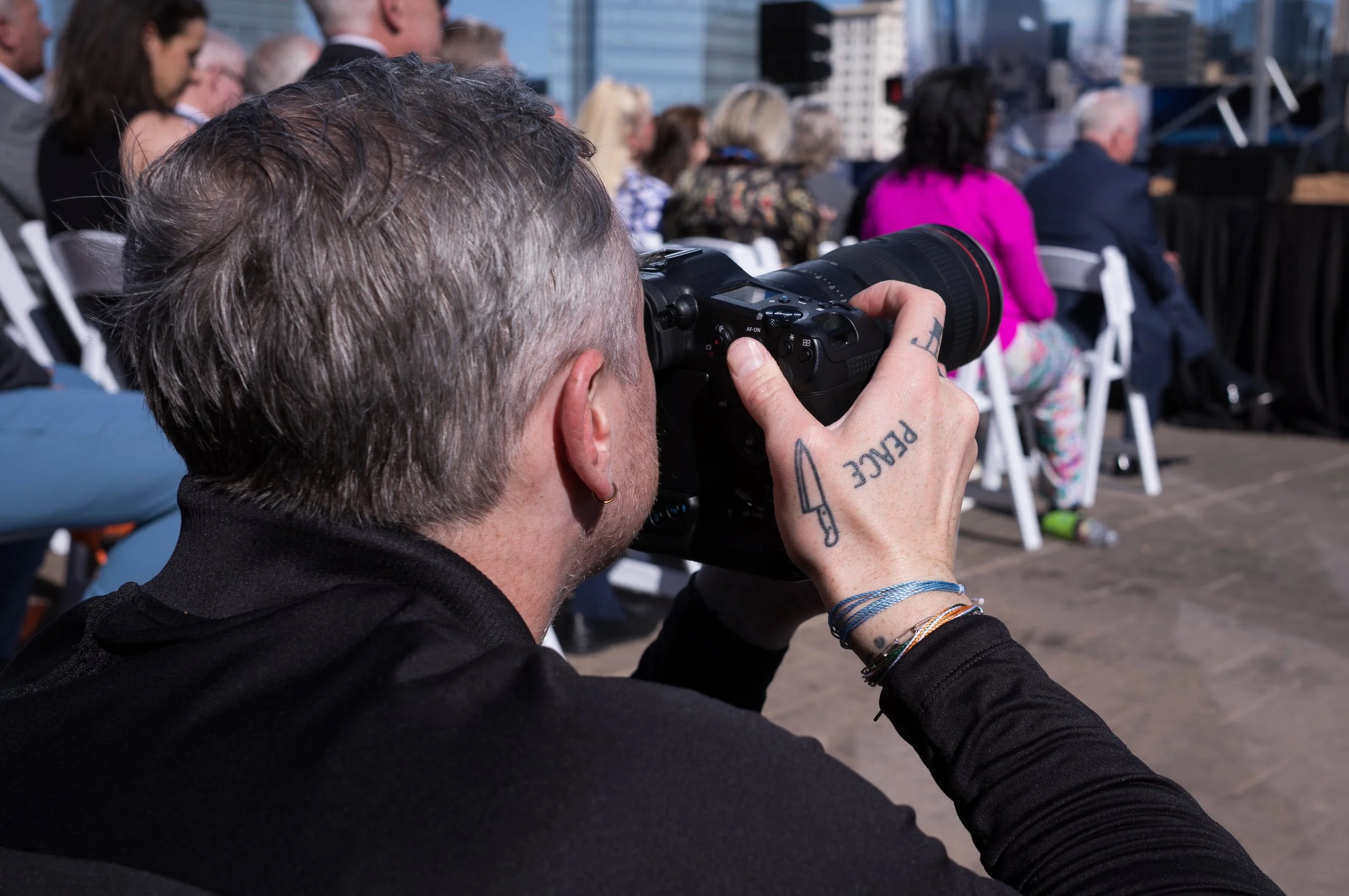 A person with tattoos on their hand holding a professional camera and taking photos at an outdoor event with a seated audience in the background.