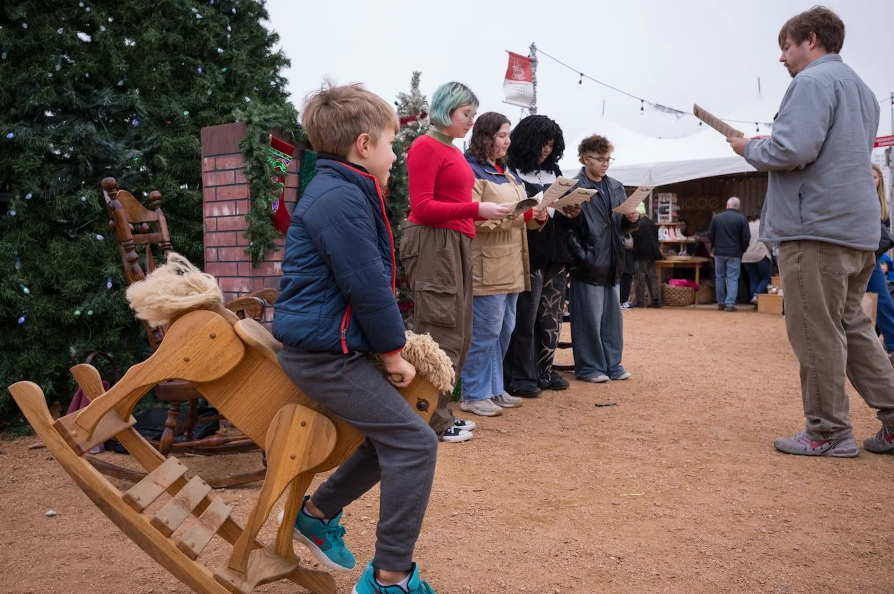 Children singing Christmas carols outdoors near a decorated Christmas tree, with one child sitting on a wooden rocking reindeer, and a person leading the group, at a holiday market.