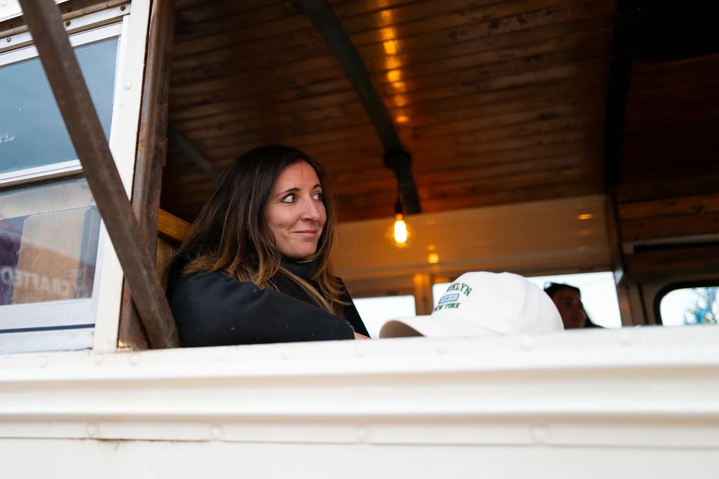 A woman with long brown hair looking out of a window in a food truck or a similar setting with wooden interior, a light bulb, and a man in the background.