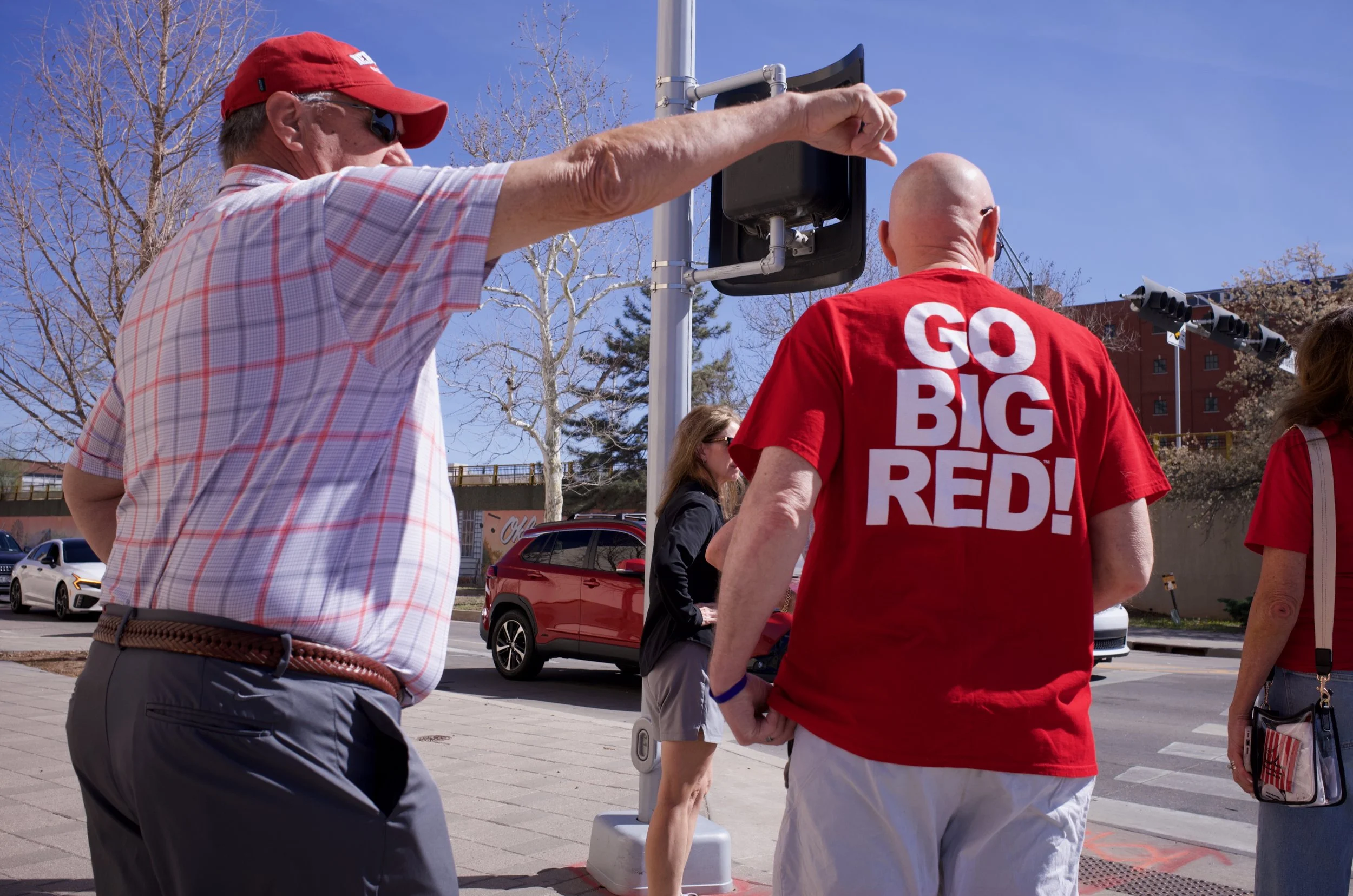 A man wearing a red cap, sunglasses, and a plaid shirt is pointing at something above. Another man with a shaved head, wearing a red T-shirt with the words "GO BIG RED!" on the back, is standing nearby. Two women are also present, one with long hair 