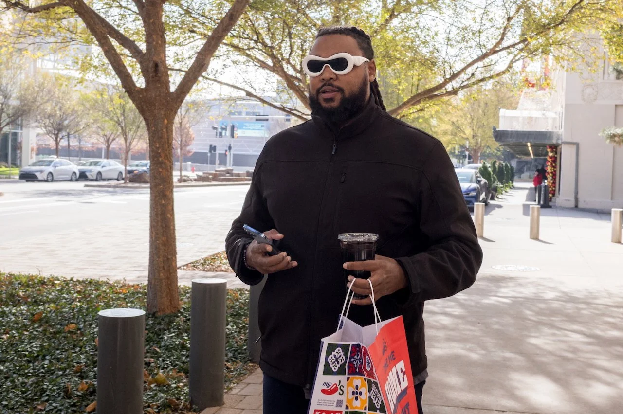 A man wearing black jacket and white sunglasses holding a Starbucks drink, a phone, and shopping bags, standing outdoors near trees and cars.