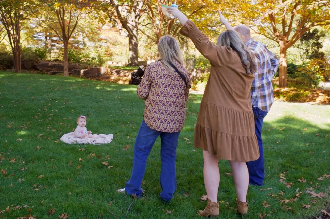 Three women taking photos of a baby girl sitting on a blanket in a park with autumn trees.