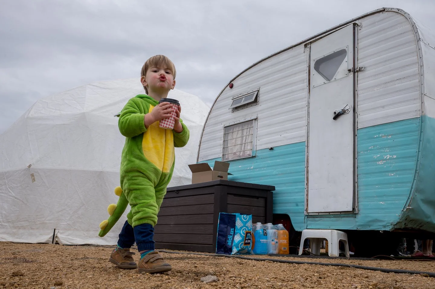 Child dressed in a dinosaur costume holding a coffee cup, standing outside near a vintage trailer and a large white tent.