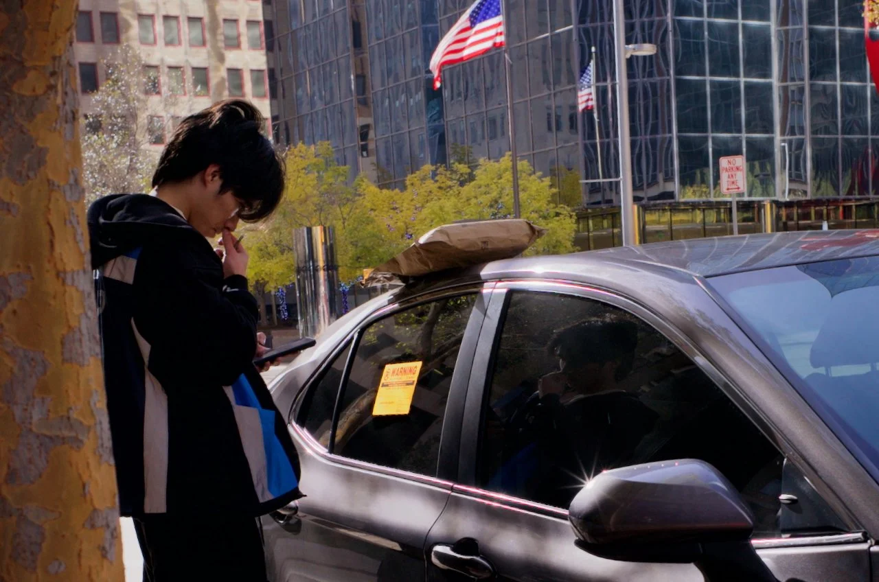 A young man is standing by a tree on a city sidewalk, looking at his phone. Behind him, there is a silver car parked with a brown paper bag on its roof. The background features tall glass buildings, American flags, and a no parking sign.