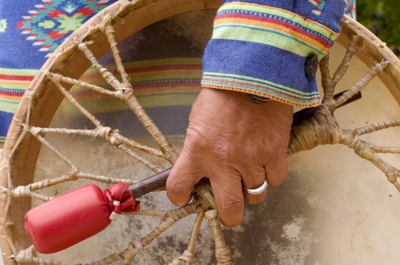 A person's hand with a silver ring on the ring finger, holding a rustic stick with a red wrapped cloth tied at the end, resting on a woven wicker basket with a colorful, striped cloth in the background.