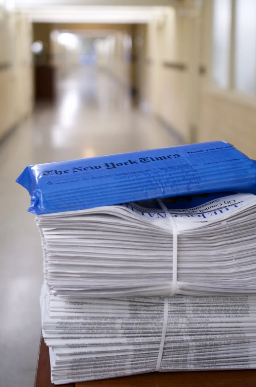 A stack of newspapers with a blue pack of The New York Times on top, placed on a wooden surface in a hallway.